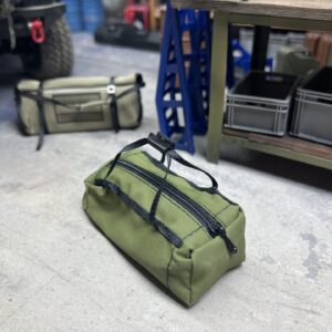 RC Scale Duffle Bag im Maßstab 1/10, olivgrün, mit funktionierendem Reißverschluss, liegend in einer realistisch gestalteten Modellbau-Werkstatt.