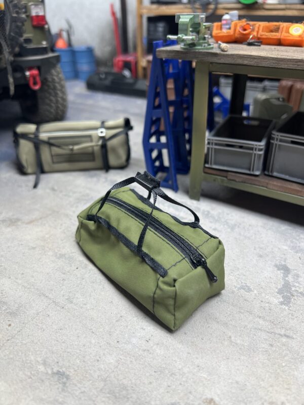RC Scale Duffle Bag im Maßstab 1/10, olivgrün, mit funktionierendem Reißverschluss, liegend in einer realistisch gestalteten Modellbau-Werkstatt.