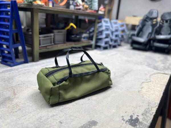 RC Scale-Duffle Bag im Maßstab 1/10, olivgrün, mit funktionierendem Reißverschluss, liegend in einer realistisch gestalteten Modellbau-Werkstatt.