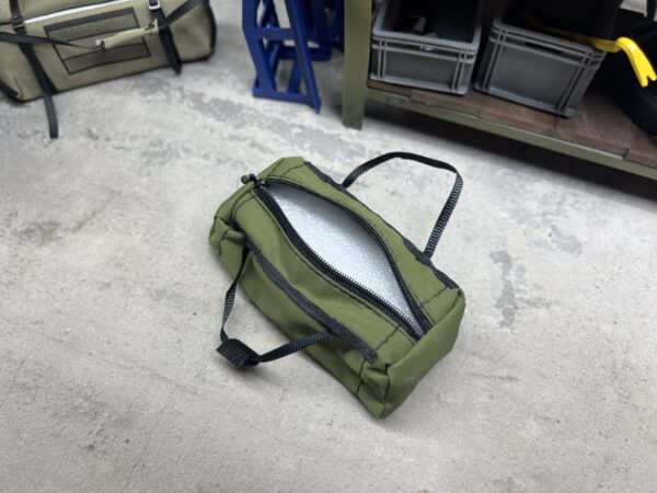 RC Scale-Duffle Bag im Maßstab 1/10, olivgrün, mit funktionierendem Reißverschluss, liegend in einer realistisch gestalteten Modellbau-Werkstatt.